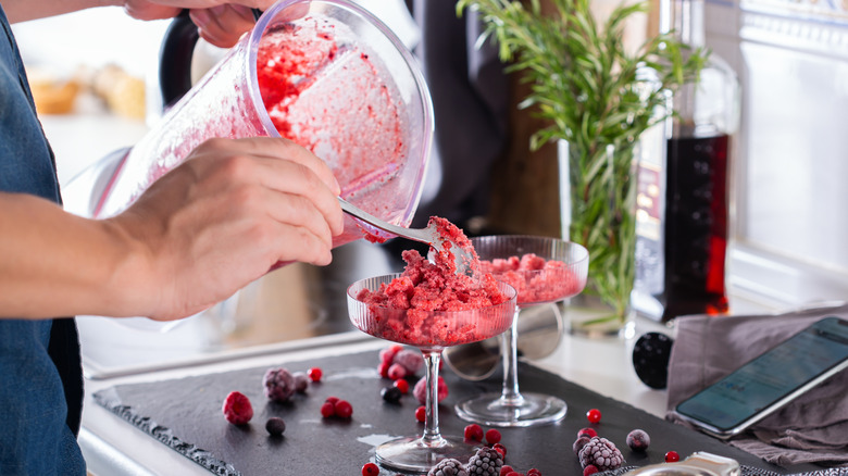 Someone scooping a blended cocktail into coupe glasses with frozen berries scattered around on the counter