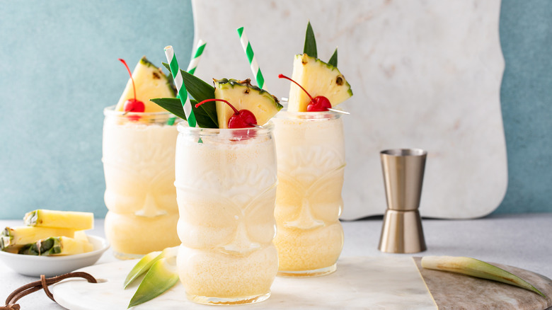 Three garnished pina coladas with a jigger and pineapple slices in the background