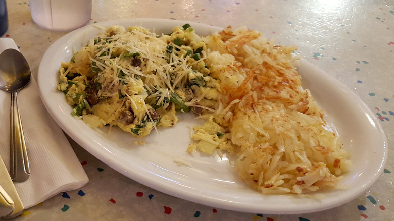 A dish with scrambled eggs, ground beef, spinach, and parmesan cheese on top with hash browns on the side and a spoon to the left