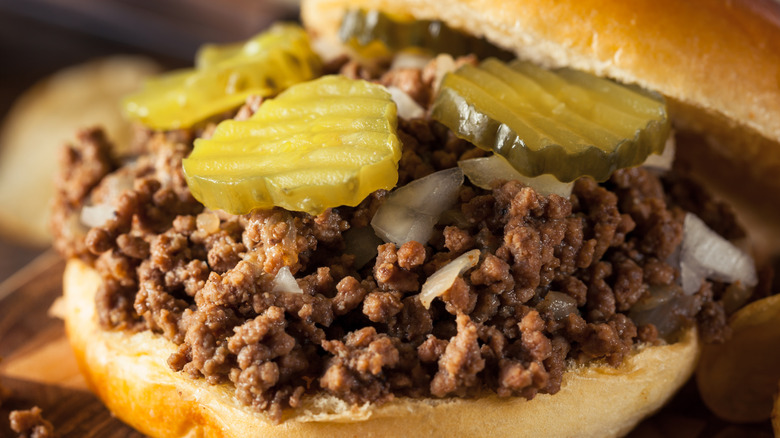 A loose meat sandwich featuring brown ground beef with onions and pickle slices on a bun