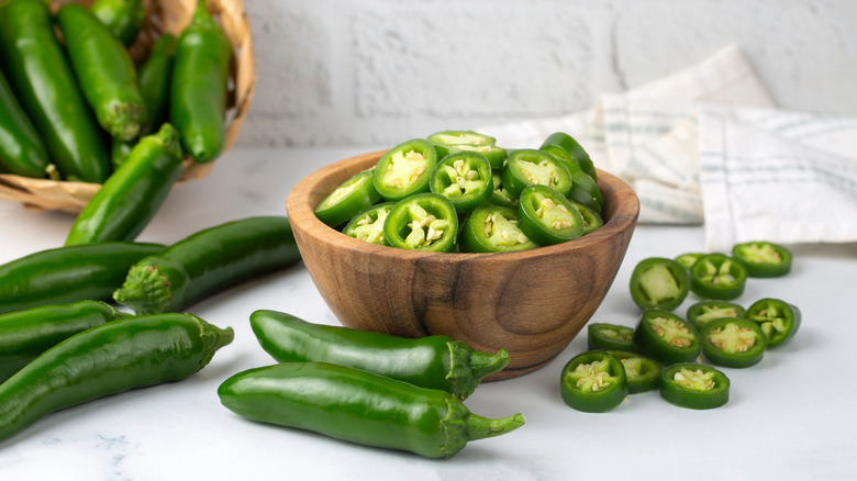 A wooden bowl full of fresh jalapeño slices on a counter, surrounded by whole and sliced chilies.