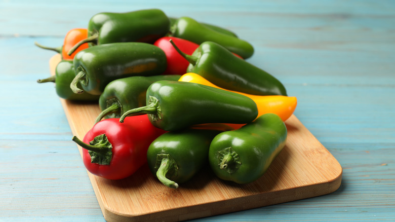 A pile of green, red, and yellow jalapeños on a wooden cutting board with blue background.
