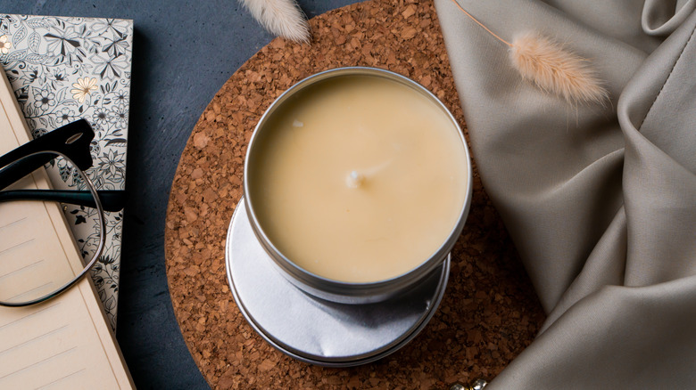 Candle in tin can on a cork mat with other items nearby, like glasses, a journal, a linen, and dried bunny tail florals.