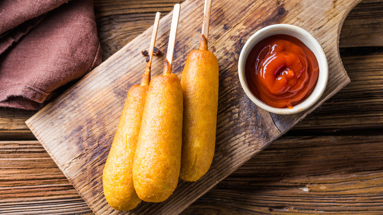 Three corn dogs on cutting board with bowl of ketchup