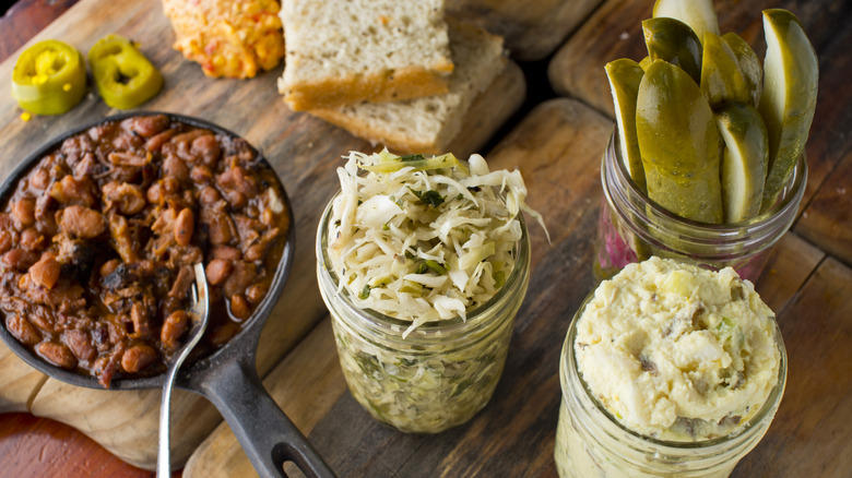 BBQ side dishes with jars of coleslaw, salad, and pickles with cast iron pan of baked beans