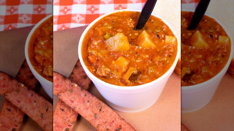 Cup of chile stew with halved sausages on red and white tablecloth