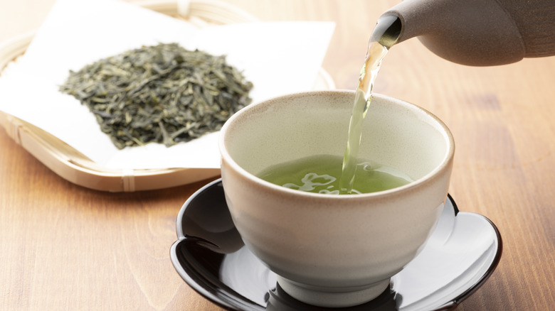 green tea being poured into a cup alongside a batch of wet green tea leaves
