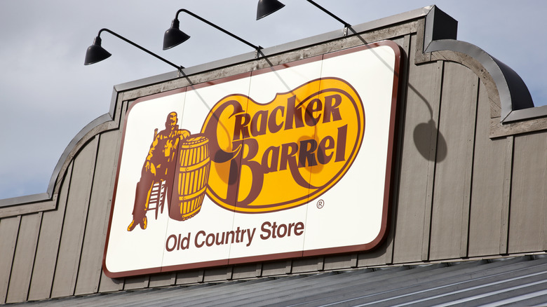 The sign on the gray roof of a Cracker Barrel, reading "Cracker Barrel Old Country Store" with an image of a man sitting in a chair next to a wooden barrel.