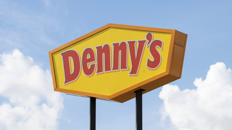 A squat, hexagonal Denny's sign displaying the restaurant's red logo on a yellow backdrop, against a blue sky with puffy white clouds.