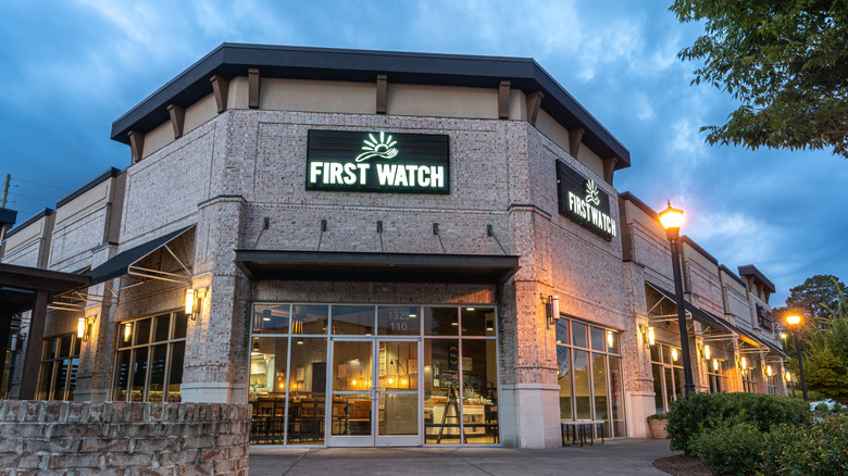 The stone exterior of a First Watch restaurant, with large glass windows and a black sign featuring an illustration of a fork and a sun, at dawn or dusk.