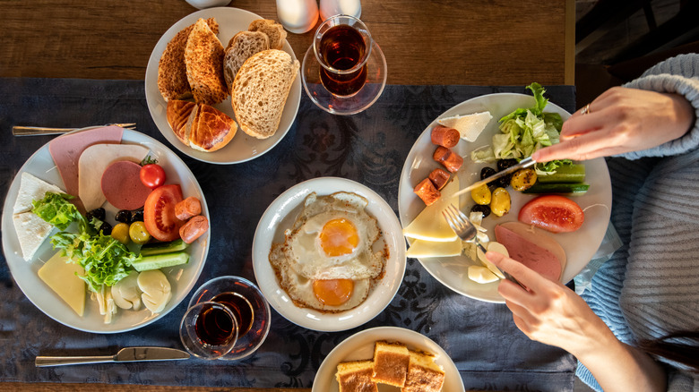 A breakfast spread including eggs, various breads, sausages, ham, and vegetables on a wooden table with a blue runner. A person in a light blue sweater cuts into a piece of cheese at the right end of the frame.