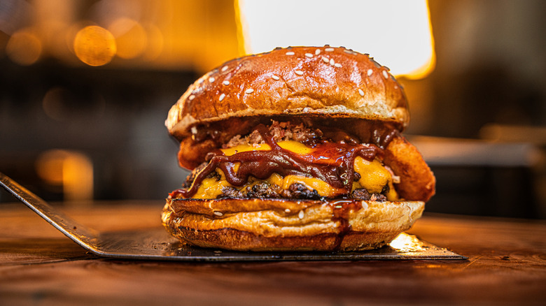 Burger with sesame seed bun featuring two melted slices of cheese, onion rings, and barbecue sauce sitting on a metal spatula.