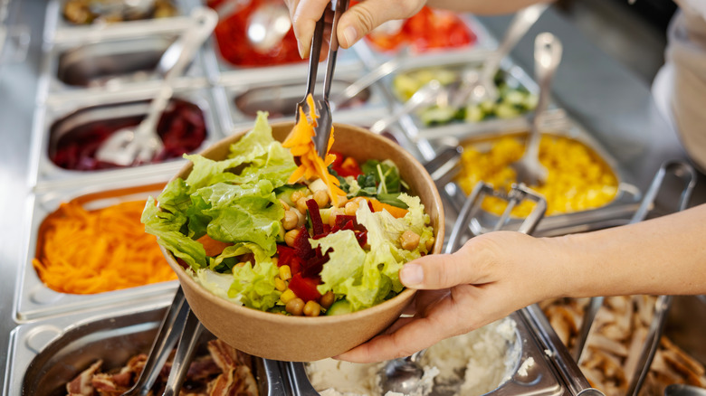 Person filling a takeaway bowl with colorful fresh ingredients from a self-service salad bar