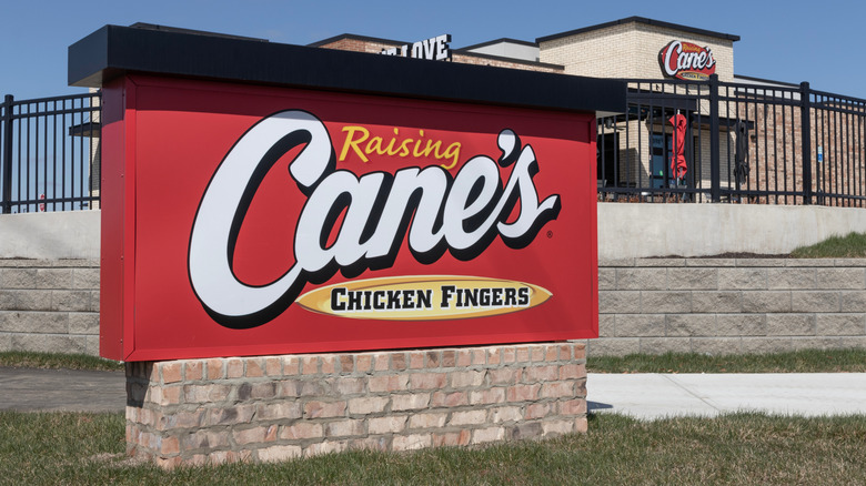 A red sign on a brick base that reads, "Raising Cane's Chicken Fingers" in a cursive font.