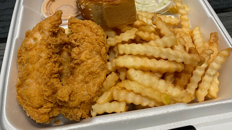 Raising Cane's golden-brown chicken fingers and crinkle-cut fries in a styrofoam takeout container. Small plastic cups of a pink sauce and coleslaw, as well as the corner of a piece of Texas Toast, are visible.
