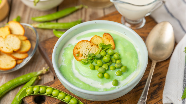 Bowl of green pea soup garnished with fresh peas, cream, and mini toasts