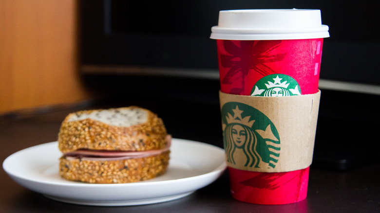 A red starbucks cup in a sleeve next to a bagel and ham sandwich on a plate