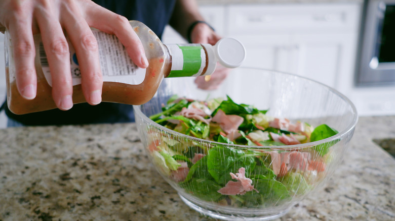 A person pouring salad dressing from a bottle onto a bowl of salad