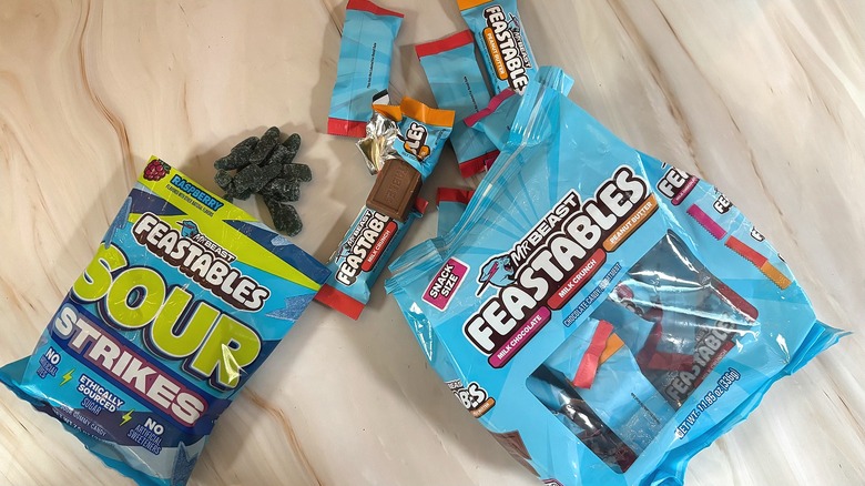 Variety bag of Feastables chocolates next to Feastables sour strikes, both spilling out of bags onto marbled surface