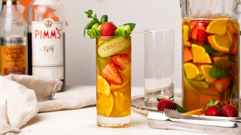 Pimm's Cup in glass with fresh fruit garnish next to pitcher and ingredient bottles in background