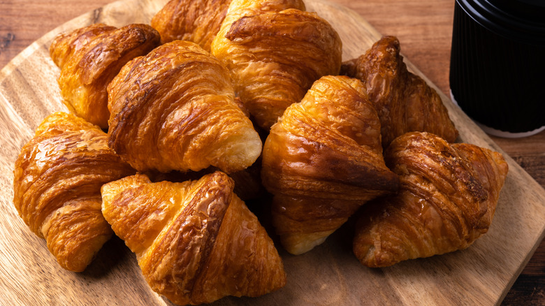 A pile of golden brown croissants sitting on a wooden cutting board, with a black paper coffee cup next to them.
