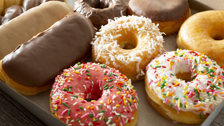 A white box containing a variety of doughnuts: classic glazed, strawberry frosted, maple bars, and Boston cream.