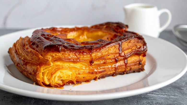 Four paper-wrapped kouign-amann pastries sit on a white plate. Their flaky layers are visible beneath a dusting of powdered sugar.