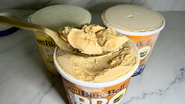 Hand holding spoonful of Blue Bell ice cream over three pints of ice cream on marble surface