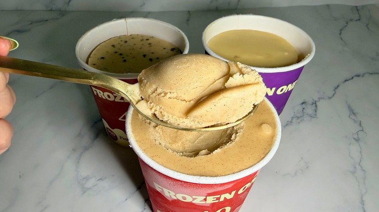 Hand holding spoonful of Frozen One ice cream over three pints of ice cream on marble surface