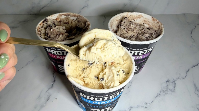 Hand holding spoonful of Protein Pints ice cream over three pints of ice cream on marble surface