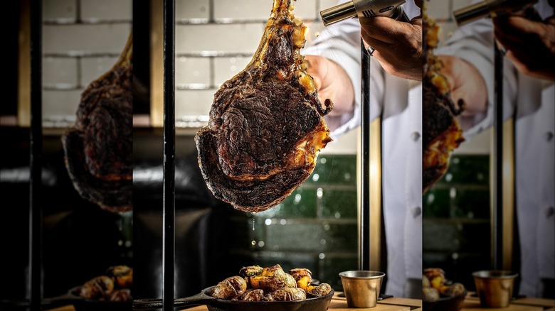 A person in a chef's uniform points a metal blowtorch at a massive tomahawk steak, hanging from a metal rack above a bowl of potatoes.
