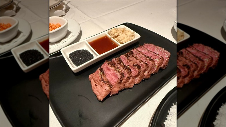 A medium rare wagyu steak lying on a black plate with seasonings and sauces in a white platter alongside it.