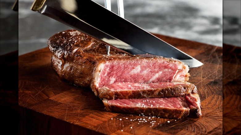 A chef knife slices through a rare Wagyu steak on top of a wooden cutting board. The steak is sparsely sprinkled with salt.