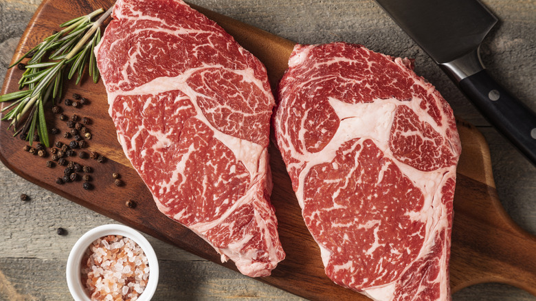 Two raw Wagyu steaks sit on a wooden cutting board, displaying intricate marbling patterns of white fat. On the side are a handful of peppercorns, two sprigs of rosemary, a ramekin of salt, and a chef knife.