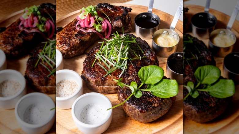 Flight of steaks on a round wooden cutting board served with salts, butter, jus, herbs, and pickled onions
