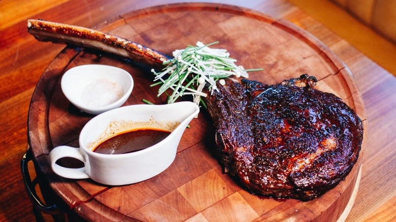 Tomahawk steak on round wooden cutting board served with greenery and sauces