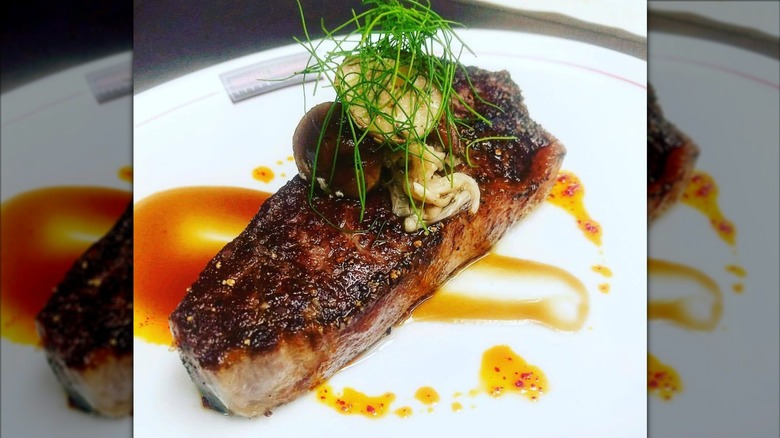 Dry-aged Wagyu NY strip on white plate topped with pickled mushrooms, greenery, and drizzled with sweet and sour plum sauce