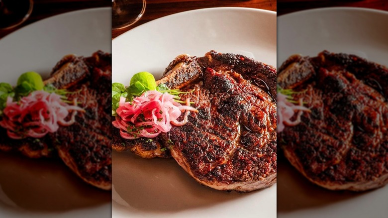 Bone-in ribeye on white plate with pickled red onions and greenery