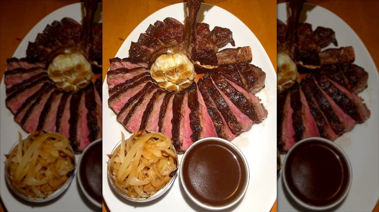 Pre-cut dry-aged porterhouse on a white plate topped with roasted garlic and served with sauteed onions and sauce
