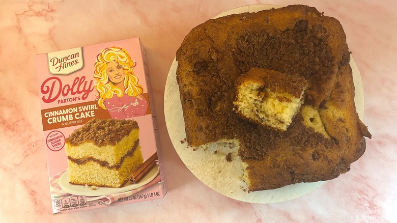 Overhead shot of box of Dolly Parton coffee cake mix next to coffee cake with slice removed on white platform