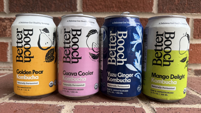 Four cans of Better Booch kombucha in different flavors lined up on brick surface