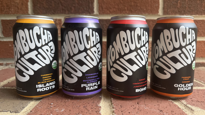 Four cans of Kombucha Culture in different flavors lined up on brick surface