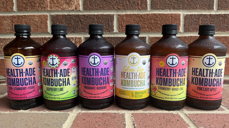 Six tinted bottles of Health-Ade kombucha lined up on brick surface