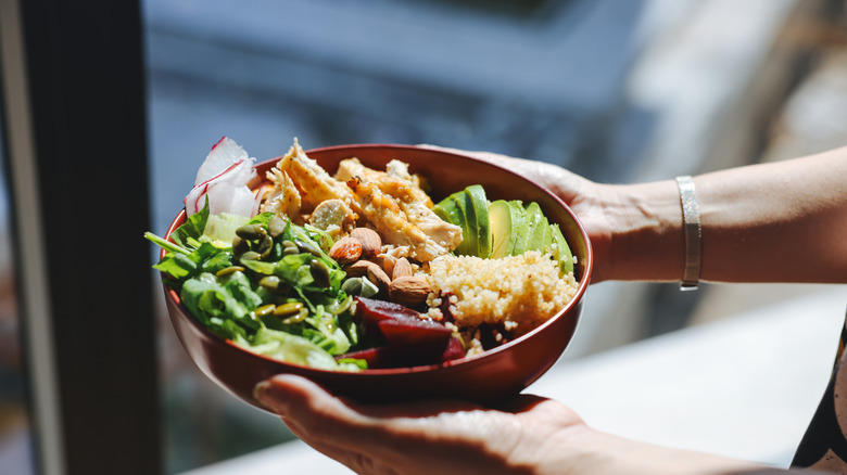 A woman's hands hold a bowl with chicken, avocado, quinoa, nuts, and greens