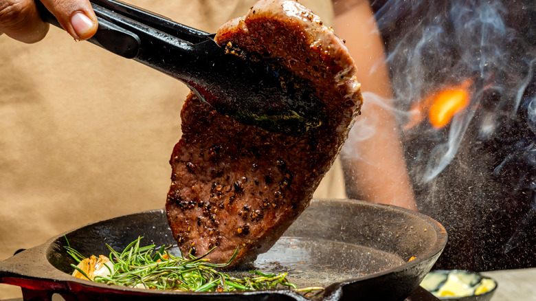 A person cooks a steak in a cast iron pan with butter and rosemary