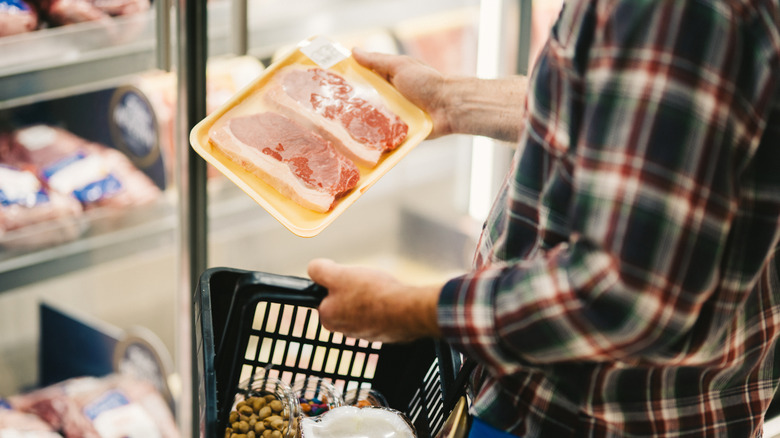 A man holding a package of beef and a green shopping basket at the grocery store