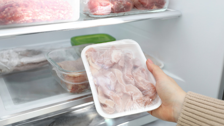 A hand holds a frozen package of chicken in front of a freezer stocked with plenty of meats