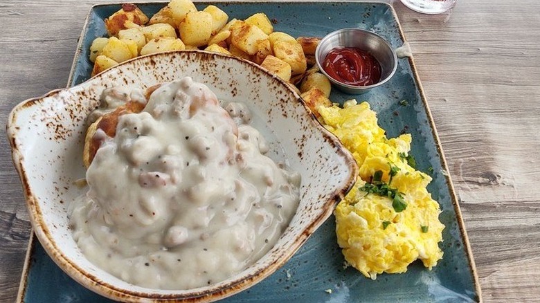 Restaurant dish at First Watch featuring biscuit covered in gravy alongside scrambled eggs and potatoes