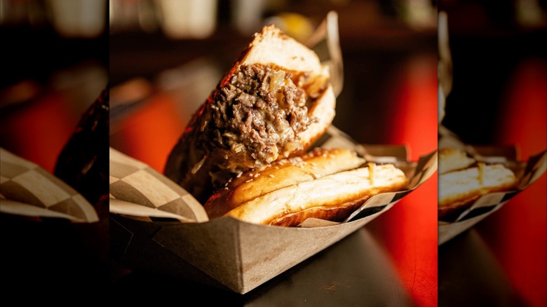 Halved cheesesteak on sesame seed roll in cardboard container