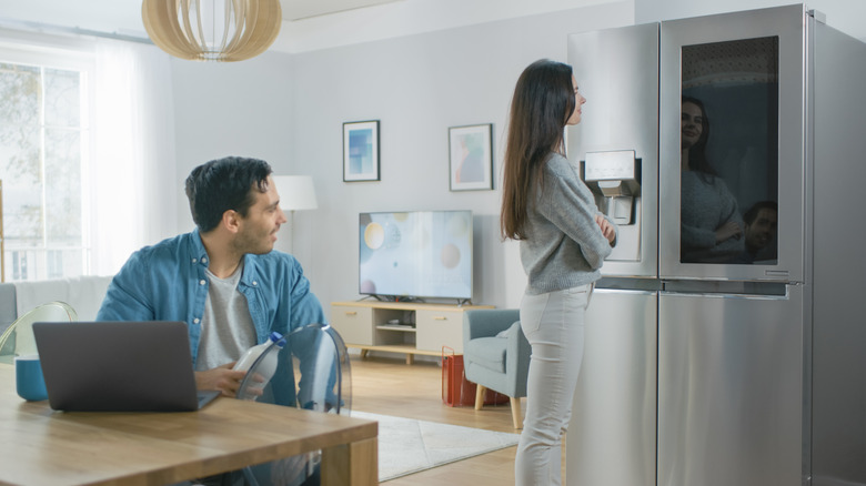 Two people in the kitchen one looking at smart fridge with the other sitting at the table
